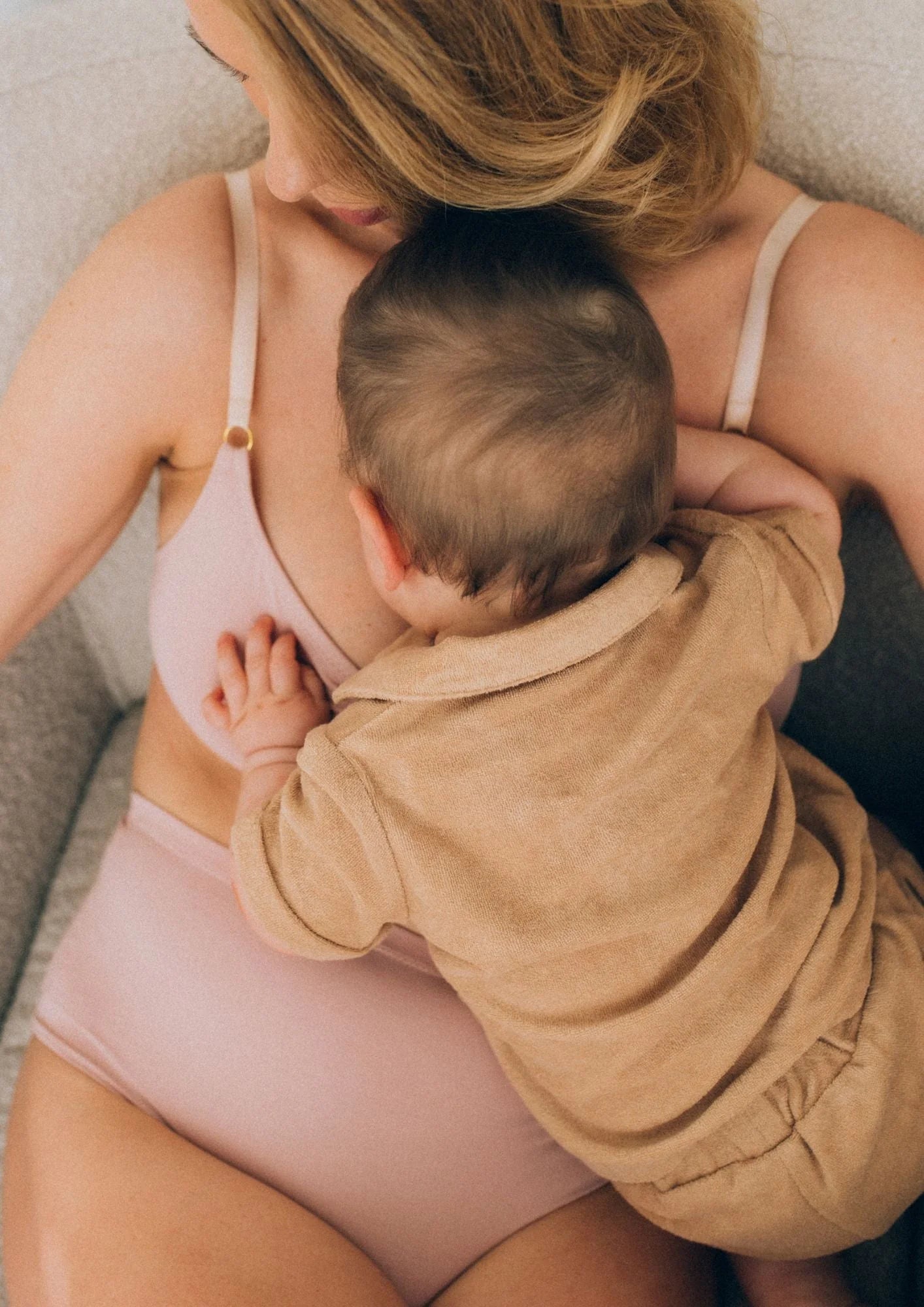 Frau sitzt auf Sessel und kuschelt mit Baby. Sie trägt rosafarbene stillfreundliche Unterwäsche.
